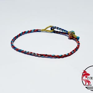 Op de afbeelding: Gevlochten armband in rood, blauw en wit. De armband heeft een goudkleurige sluiting en een decoratieve knoop met een kleine blauwe kraal. Het Amori Collective-logo staat rechtsonder.
