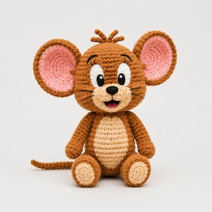 Puede incluir: Un peluche de ganchillo con forma de ratón, cuerpo marrón y vientre y almohadillas de color beige claro. El ratón tiene grandes orejas redondas con centros rosas, ojos bordados en negro, una nariz negra y una larga cola hacia la izquierda.