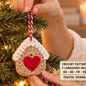 Pode incluir: Um ornamento de crochê em forma de casa na cor bege com um telhado branco e um coração vermelho no centro, suspenso por um cordão trançado vermelho e branco. Uma caixa de texto diz: CROCHET PATTERN PDF 5 LANGUAGES INCLUDED EN • DE • FR • ES • PL DIGITAL DOWNLOAD.