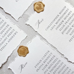 May include: Close-up of elegant white place cards with torn edges, featuring guest names in black script. Each card is adorned with a gold wax seal. The cards are arranged on a neutral surface, ideal for weddings or special events.
