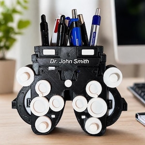 Puede incluir: Un portalápices negro y blanco impreso en 3D con forma de máquina de examen ocular. El soporte está personalizado con el texto "Dr. John Smith" y contiene varios bolígrafos. El diseño presenta detalles circulares blancos y un área rectangular para guardar bolígrafos.