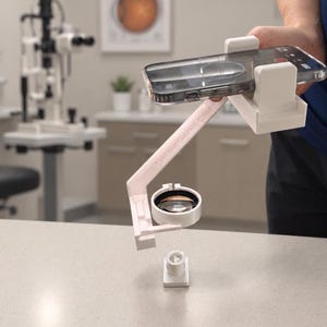 Puede incluir: Un adaptador para smartphone EyePrint Fundus Cam en color blanco que incluye una base para dispositivo móvil, un brazo de soporte en ángulo y una carcasa circular para lentes. El dispositivo alinea la cámara con una lente óptica.