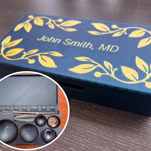 Könnte beinhalten: Eine marineblaue Box mit goldenem Blattdesign und dem Text "John Smith, MD". Die Box ist geöffnet und zeigt verschiedene medizinische Werkzeuge und Behälter. Die Box steht auf einer Holzoberfläche.