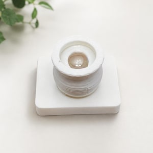 May include: A white cylindrical object with a wide, flat rim and a smooth, rounded interior basin, mounted on a square white base. The object features a layered, ribbed texture along its exterior sides and a glossy, light beige centre.