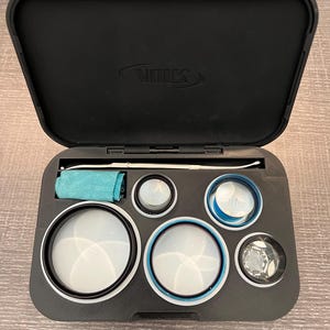 May include: A black case with a VOLK logo on the lid, containing a set of lens filters and a cleaning cloth. The filters are various sizes and have blue and white markings.