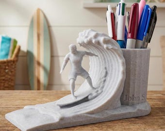 May include: A grey pen holder depicting a surfer on a wave. The holder includes a cylindrical section for pens and pencils, and a base with the name "John S" engraved. Several pens and markers are visible inside the holder.