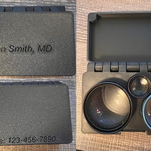 May include: A dark grey, rectangular case with the name "John Smith, MD" and phone number "123-456-7890" engraved on it. The case opens to reveal three lenses of varying sizes. The lenses are black with a blue ring around one.