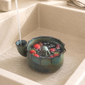 Può includere: Uno scolabacche in ceramica rotondo, color verde acqua e blu scuro, con bordo smerlato e una piccola maniglia laterale. Lo scolabacche è riempito di fragole e mirtilli freschi, immersi nell'acqua, con un foro di scarico centrale visibile.