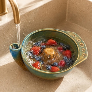 Puede incluir: Un cuenco colador de cerámica redondo, en tonos verde azulado y marrón, lleno de agua, fresas frescas y arándanos. El cuenco cuenta con una cúpula de drenaje central, un pico lateral para el agua y un asa con cuatro orificios circulares.