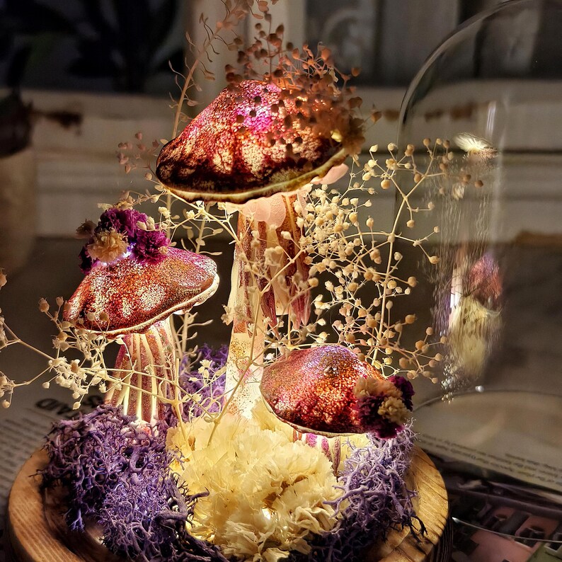 Puede incluir: Un terrario decorativo de hongos con tres hongos texturizados iluminados en tonos rojos y dorados. Los hongos est&aacute;n rodeados de flores secas, musgo morado y una campana de cristal. La base es un cuenco de madera.