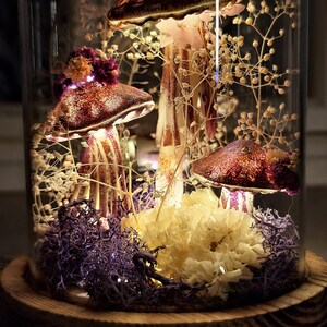 Puede incluir: Una campana de cristal decorativa con esculturas de hongos iluminadas. Los hongos tienen sombreros marrones con motas blancas y tallos rayados. La base incluye musgo morado, flores amarillas y delicadas ramitas blancas, todo iluminado por peque&ntilde;as luces de hadas.