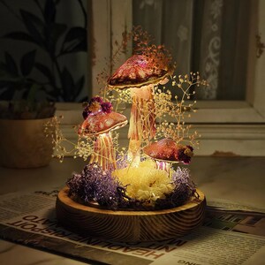 Puede incluir: Un terrario decorativo de hongos con tres sombreros de hongos iluminados de color marr&oacute;n rojizo. Los hongos est&aacute;n rodeados de flores secas y musgo morado, colocados sobre una base de madera. El terrario est&aacute; iluminado desde dentro, creando un brillo c&aacute;lido.