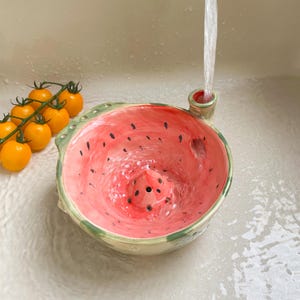 May include: A ceramic colander shaped like a halved watermelon with a pink interior, black seeds, and a green rind. Water flows from a faucet into the bowl, which features drainage holes at the bottom and side. A cluster of yellow cherry tomatoes sits nearby.
