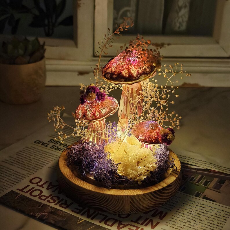 Puede incluir: Un terrario decorativo de setas con tres sombreros de setas rojos y rosas brillantes iluminados. Las setas est&aacute;n rodeadas de flores secas y musgo, colocadas en una base de madera. El terrario est&aacute; iluminado desde dentro.