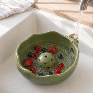 May include: A round, olive green ceramic berry colander with a central perforated dome and a side spout. The bowl is filled with water and fresh strawberries and blueberries, with a stream of water flowing from a faucet into the spout.