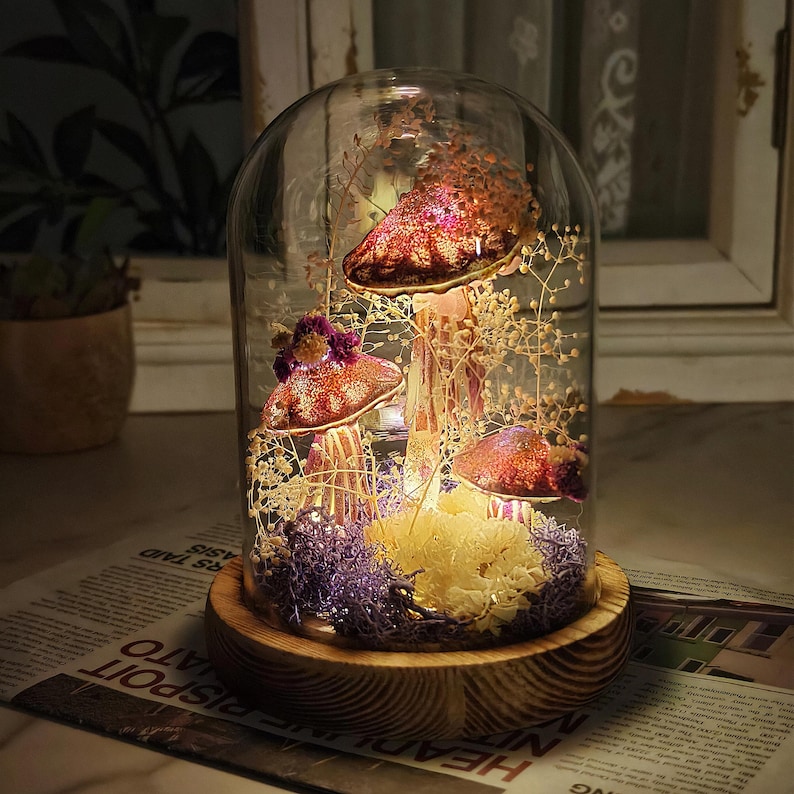 Puede incluir: Una campana de cristal decorativa con setas artificiales iluminadas. Las setas tienen sombreros rojos y dorados con purpurina y est&aacute;n rodeadas de flores secas y musgo. La base es de madera.