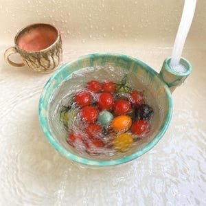Pode incluir: Uma tigela escorredor de cerâmica turquesa com alça lateral e bico integrado, cheia de água e tomates cereja variados. Uma caneca de cerâmica turquesa e rosa a condizer está por perto, enquanto a água flui para o bico do escorredor.