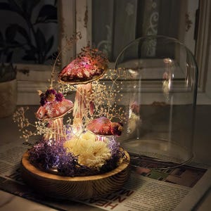 Puede incluir: Una exhibici&oacute;n decorativa de hongos con tres sombreros iluminados cubiertos de purpurina en tonos rojos y rosas. Los hongos est&aacute;n colocados sobre una base de madera con musgo morado y flores secas, bajo una campana de cristal. Luces de hadas a&ntilde;aden un brillo c&aacute;lido.
