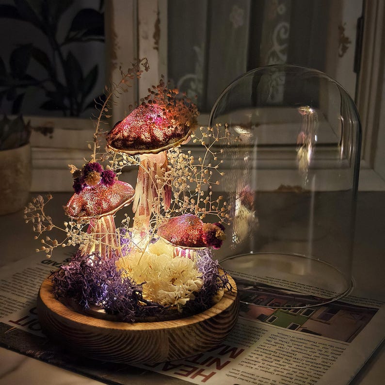 Puede incluir: Una exhibici&oacute;n decorativa de hongos con tres sombreros iluminados cubiertos de purpurina en tonos rojos y rosas. Los hongos est&aacute;n colocados sobre una base de madera con musgo morado y flores secas, bajo una campana de cristal. Luces de hadas a&ntilde;aden un brillo c&aacute;lido.