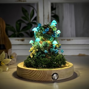 Puede incluir: Lámpara decorativa con forma de hongo y base de madera. La lámpara presenta hongos azules brillantes en musgo verde, con flores azules y amarillas. La base de madera tiene un botón plateado.