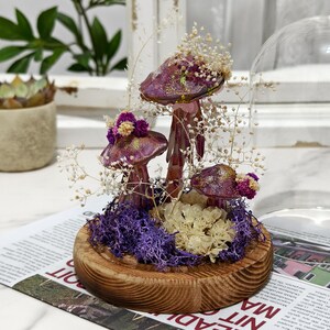Puede incluir: Una campana de cristal decorativa con un arreglo de setas caprichoso. Las setas tienen sombreros morados con detalles dorados y est&aacute;n rodeadas de flores secas y musgo morado, sobre una base de madera.