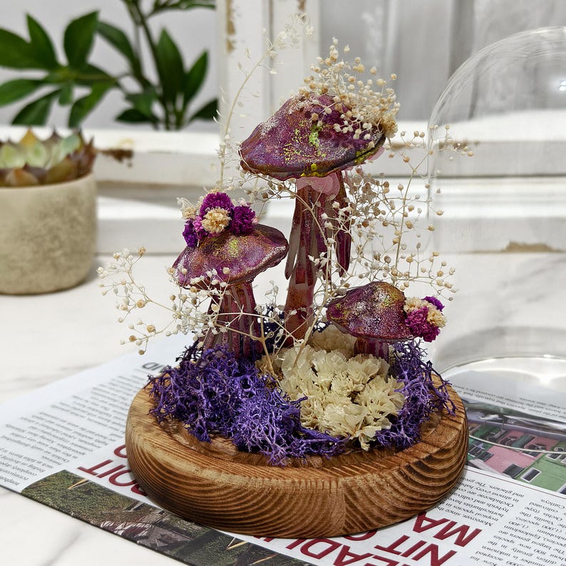 Puede incluir: Una campana de cristal decorativa con un arreglo de setas caprichoso. Las setas tienen sombreros morados con detalles dorados y est&aacute;n rodeadas de flores secas y musgo morado, sobre una base de madera.