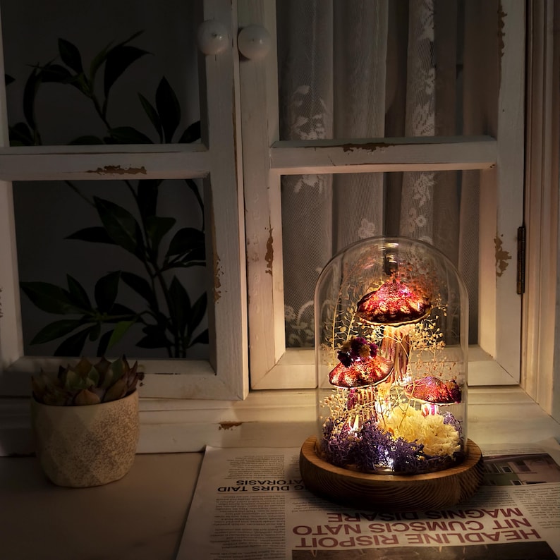 Puede incluir: Una campana de cristal decorativa con tres figuras de setas rojas y doradas, iluminadas por peque&ntilde;as luces. Las setas est&aacute;n rodeadas de flores secas y se asientan sobre una base de madera. La campana est&aacute; cerca de una ventana.