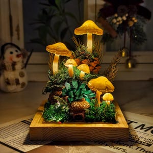 Könnte beinhalten: Ein dekoratives, beleuchtetes Pilz-Diorama. Die Szene zeigt mehrere leuchtende gelbe Pilze unterschiedlicher Größe, eingebettet in grünes Moos, Tannenzapfen und kleine Pflanzen, alles in einem Holzrahmen. Ein kleines Haus ist zwischen den Pflanzen eingebettet.