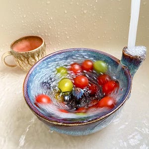 May include: A blue and brown ceramic colander bowl with a side handle and integrated water spout, filled with red and green cherry tomatoes being rinsed under a stream of water, alongside a matching textured ceramic mug with a light brown finish.