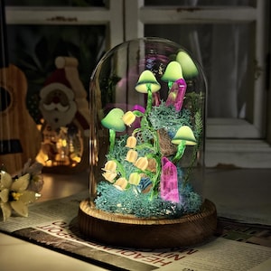 Puede incluir: Una campana de cristal decorativa con base de madera, que muestra una escena en miniatura iluminada de hongos y cristales. Los hongos tienen sombreros verdes y están iluminados con un brillo suave. Los cristales rosas y el musgo añaden color y textura.
