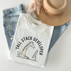 Peut inclure: T-shirt blanc à col rond avec un motif de girafe et le texte "TALL STACK DEVELOPER" en noir. La girafe est représentée assise devant un écran d'ordinateur. Le t-shirt est posé sur un jean bleu, un chapeau et des fleurs séchées.