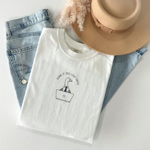 Peut inclure: T-shirt blanc à col rond avec un dessin d'oie de dessin animé travaillant sur un ordinateur portable, avec le texte "Honk if you love data". Le t-shirt est associé à un jean bleu et un chapeau beige.