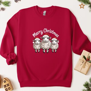 May include: A red Christmas sweatshirt with a graphic of three sheep wearing Santa hats. The text "Merry Christmas" is above the sheep. The image is decorated with festive elements.