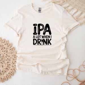Può includere: Una maglietta bianca con testo nero che recita "IPA A LOT WHEN I DRINK" con un'immagine di un bicchiere di birra.