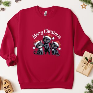May include: A red sweatshirt with a festive design. It features three black cats in Santa hats, with the words "Merry Christmas" above them. The design is set against a backdrop of Christmas decorations.