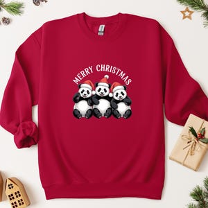 May include: A red sweatshirt with a festive design. The sweatshirt features three pandas wearing Santa hats, with the words "MERRY CHRISTMAS" above them in white. A seasonal, holiday-themed garment.