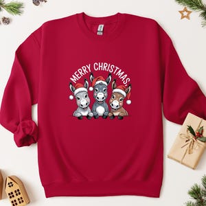 May include: A red sweatshirt with a graphic of three donkeys wearing Santa hats and the text "MERRY CHRISTMAS" above them. The donkeys are in shades of grey and brown. A festive Christmas jumper for the holiday season.