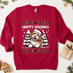 Könnte beinhalten: Ein rotes Sweatshirt mit festlichem Design. Die Grafik zeigt einen Hund mit Weihnachtsmütze, schneebedeckte Bäume und den Text "HAPPY DOGMAS". Das Design umfasst Lebkuchenmänner, Zuckerstangen und Schneeflocken.