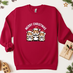 May include: A red sweatshirt with a graphic of three hamsters wearing Santa hats, and the words "Merry Christmas". A festive item for the Christmas holiday season.