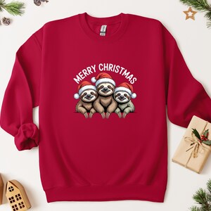 May include: A red sweatshirt with a graphic of three sloths wearing Santa hats and the text "MERRY CHRISTMAS". The sloths are brown and grey, and the hats are red and white. A festive item for the Christmas holiday season.