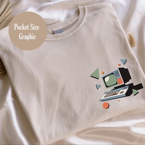 Puede incluir: Camiseta beige con un gráfico de ordenador retro. El diseño presenta un ordenador vintage con una pantalla verde, teclado y formas geométricas coloridas. El texto "Pocket Size Graphic" está impreso en la camiseta.