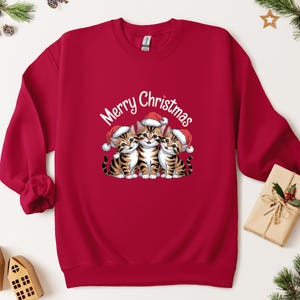 May include: A red sweatshirt with a festive design featuring three tabby kittens wearing Santa hats. The words "Merry Christmas" are written above the kittens in a white script. A seasonal holiday garment.
