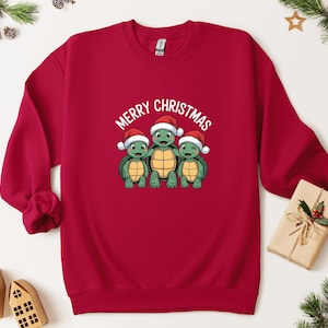 May include: A red crewneck sweatshirt featuring a graphic of three cartoon turtles wearing Santa hats, with the text "Merry Christmas" above them. The turtles have green bodies and yellow shells. The sweatshirt is a solid red color.