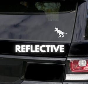 May include: White reflective sticker with a T-Rex silhouette and the word "REFLECTIVE" in capital letters. The sticker is designed for a vehicle's rear window or body panel. The image is set against a black background.