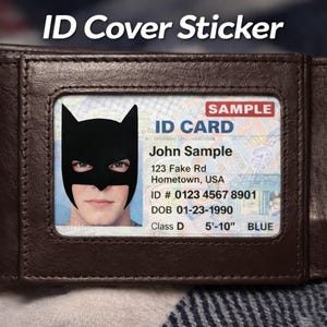Könnte beinhalten: Ein braunes Lederportemonnaie mit einem sichtbaren Ausweis. Der Ausweis zeigt eine schwarze Batman-Maske über dem Gesicht einer Person. Der Text "ID Cover Sticker" steht oben.