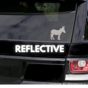 Op de afbeelding: Een zwarte auto met een wit silhouet van een ezel en het woord "REFLECTIVE" in witte letters op de achterkant.