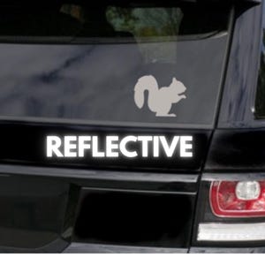 May include: A reflective car decal featuring a silhouette of a squirrel. Below the squirrel is the word "REFLECTIVE" in bold, white capital letters. The decal is designed to be applied to a car window or surface.