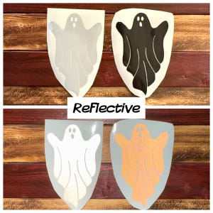 Puede incluir: Cuatro pegatinas en forma de fantasma, dos blancas, una negra y una naranja, con acabado reflectante. Las pegatinas están sobre un fondo de madera. La palabra "Reflectivo" está impresa debajo de las pegatinas.