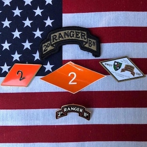 Puede incluir: Una colección de parches y pines militares sobre una bandera estadounidense roja, blanca y azul. Los parches incluyen un parche 2nd Ranger Bn, un pin 2nd Ranger Bn y un parche GWOT. Los pines incluyen un pin en forma de diamante con el número 2 en él y un pin rectangular con el número 2 en él.