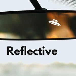 Puede incluir: Un espejo retrovisor negro con una pegatina de OVNI blanca en el cristal. El texto "Reflective" está impreso debajo del espejo.